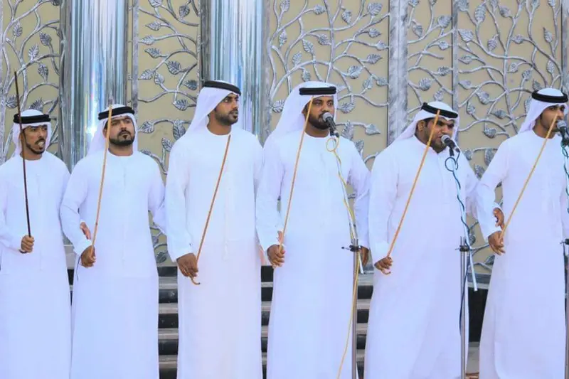 Ayala troupe in white kandura holding thin canes and chanting at microphones at an event in the UAE